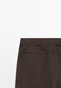 Pantalon marron foncé avec taille élastique et deux poches passepoilées horizontales à l'arrière, vu de dos sur fond blanc.