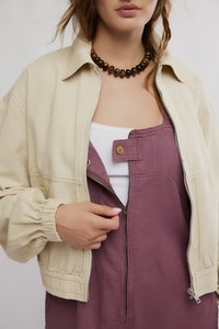 Beige Bomberjacke mit gerafften Ärmeln, getragen über einem lila Kleid mit Frontreißverschluss, darunter ein weißes Tanktop und eine Perlenkette.