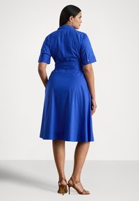 Ženska stoji, oblečena v modro midi obleko s kratkimi rokavi in pasom ter nosi rjave sandale s peto, obrnjena stran proti enobarvnemu ozadju.