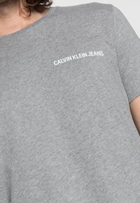 Šedé tričko vyrobené z bavlny s kulatým výstřihem a krátkými rukávy. Na levé straně hrudi se nachází bílý nápis "CALVIN KLEIN JEANS".