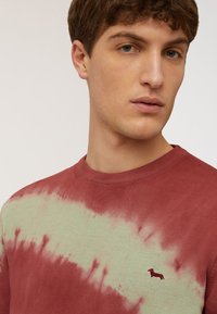 Rood en groen tie-dye sweatshirt met geribbelde kraag; heeft een klein geborduurd hondenlogo op de borst. Zachte textuur en casual ontwerp.