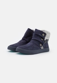 Botas de ante azul marino hasta el tobillo con puños doblados, tres botones decorativos, forro de pelo y suelas planas de goma sobre un fondo blanco.