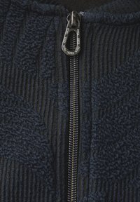 Chaqueta de tela texturizada azul marino con cuello alto, que cuenta con un cierre metálico de longitud completa. Los patrones acanalados y lisos crean un contraste visual.