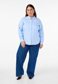 Camicia a maniche lunghe azzurro chiaro con colletto, apertura frontale con bottoni. Abbinata a jeans in denim blu scuro a gamba larga. Tessuto dalla superficie liscia.