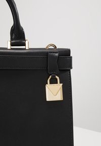 Borsa a tracolla in pelle nera con manico superiore e dettaglio di lucchetto dorato appeso a una tracolla vicino al bordo superiore.
