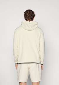 Lys beige hættebluse med en glat tekstur, der har sorte accenter ved kanten og manchetterne. Set fra ryggen.