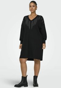 Abito nero oversize con maniche lunghe, scollo a V e applicazioni di borchie sulle spalle. Abbinato a stivaletti neri alla caviglia.