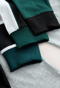 Gros plan sur des manches de sweat-shirt pliées avec des poignets côtelés en noir, vert et blanc, reposant sur un fond en tissu gris clair.