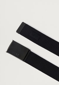 Crni tkani najlonski pojas s pravokutnom crnom metalnom kopčom. Ima uzdignuti logo na kopči i zakrpu na kraju pojasa.