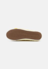 Sneaker sole in brown rubber with wavy patterns, beige edges, and yellow accents. Features textured grip design and embossed logo.