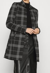 Černobílý kostkovaný blazer se strukturovaným designem, dlouhými rukávy a decentním límcem. Spárován s černou koženou sukní.