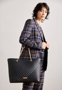 Black quilted tote bag with diagonal stitching, gold chain handles, and gold logo detail. Model wears a brown and blue plaid suit.