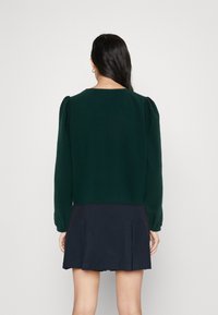 Dark green long-sleeve top with puffed shoulders, textured fabric, and a round neckline paired with a navy pleated skirt.