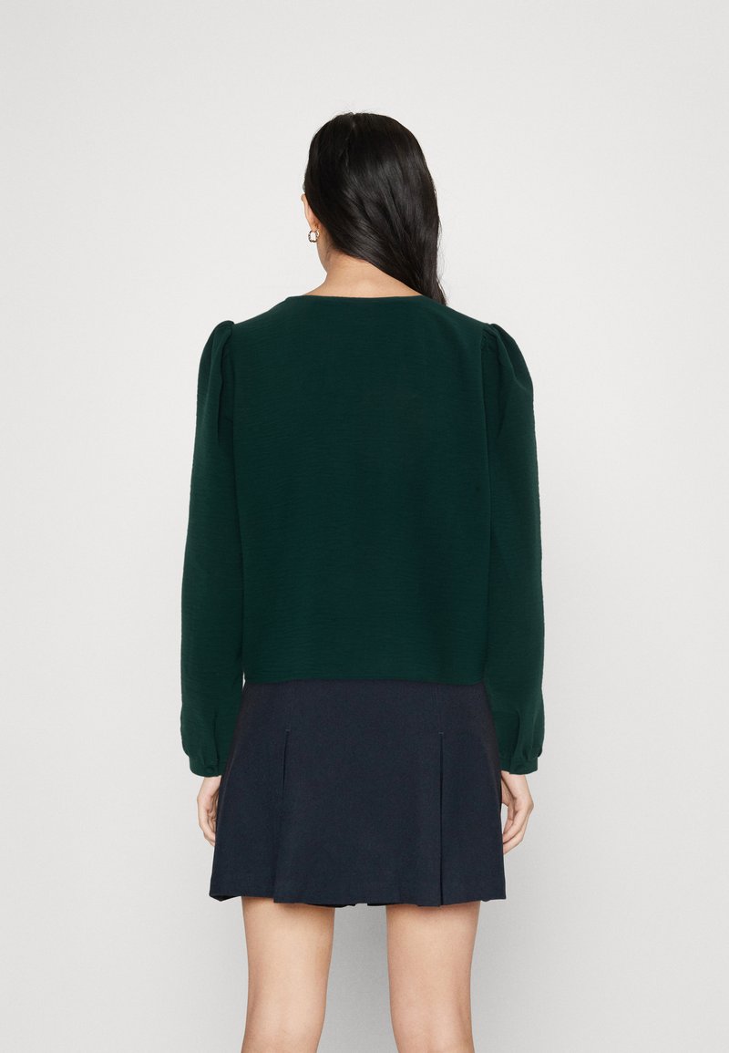 Dunkelgrünes Langarmoberteil mit gepufften Schultern, strukturiertem Stoff und rundem Halsausschnitt, kombiniert mit einem marineblauen Faltenrock.