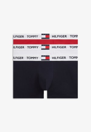 Homme musclé debout, pieds nus, bras croisés, portant un boxer rouge Tommy Hilfiger sur fond blanc uni.