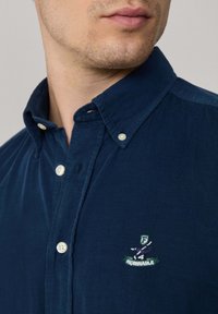 Navyblauwe corduroy knoopsluiting shirt met een klassieke kraag en geborduurd logo op de linkerborst. Zachte textuur met kleine knopen.