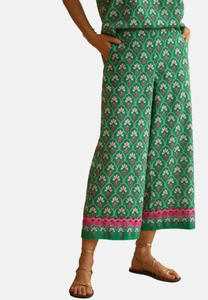 Groene wijde broek met roze bloemenpatroon en roze bies aan de zoom, gedragen met gouden platte sandalen met bandjes en handen in de zakken.