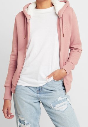 Persona che indossa una felpa leggera rosa con zip e fodera in pile bianca, t-shirt bianca e jeans strappati azzurri, in piedi con una mano in tasca.