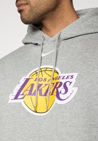 Sweat à capuche gris avec une poche kangourou, arborant un grand logo des Los Angeles Lakers en jaune et violet, agrémenté de cordons argentés.