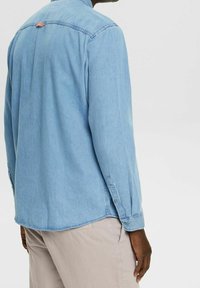 Camisa de mezclilla azul claro con mangas largas, puños con botones y dobladillo redondeado. Presenta un pequeño detalle de asa en la parte trasera.