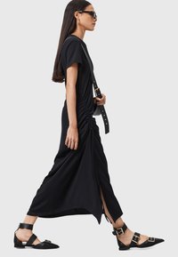 Abito midi nero con arricciature laterali e spacco, caratterizzato da maniche corte. Abbinato a ballerine nere con punta appuntita, con cinturini regolabili e dettagli dorati.
