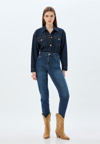 Chaqueta de mezclilla oscura con cierre de botones y dos bolsillos en el pecho; combinada con jeans de cintura alta en azul oscuro y botas de cowboy en color marrón.