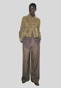 Veste en daim olive avec des boutons-pression, superposée à une chemise marron, assortie à un pantalon marron ample. Tissu texturé et coupe décontractée.
