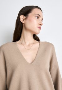 Beige V-Ausschnitt-Pullover mit weiten Ärmeln, aus weichem Strickmaterial hergestellt. Er hat eine subtile Textur und eine zarte goldene Halskette mit einem Kreuzanhänger.