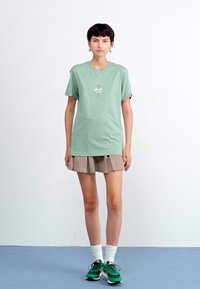 Grön bomullströja med korta ärmar och en liten broderad design på bröstet, ihop med en beige veckad kjol och gröna sneakers.