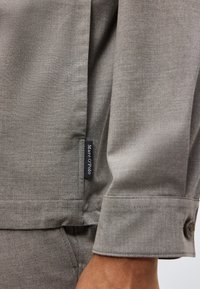 Nærbillede af manchet og skørt på en grå, langærmet skjorte med et sort "Marc O'Polo"-mærke på sidesømmen.