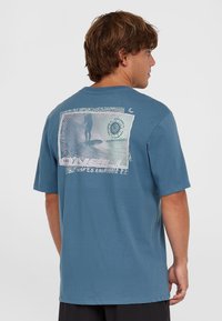 Camiseta de algodón azul con mangas cortas, que presenta una impresión gráfica de un surfista y un diseño en espiral en la parte posterior. Corte relajado.