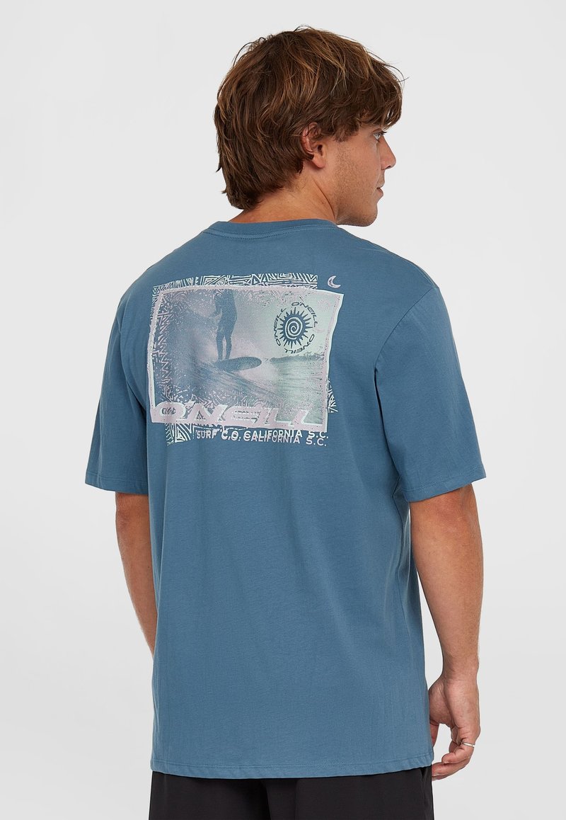 Camiseta de algodón azul con mangas cortas, que presenta una impresión gráfica de un surfista y un diseño en espiral en la parte posterior. Corte relajado.