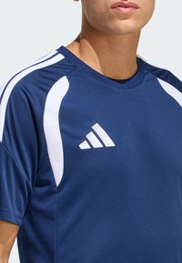 Μπλε σκούρο αθλητικό μπλουζάκι κατασκευασμένο από υφασμάτινη υφή, με λευκές λεπτομέρειες στους ώμους και λευκό λογότυπο Adidas στο στήθος. Στρογγυλή λαιμόκοψη.