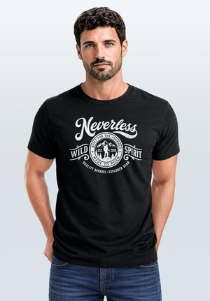 Mann mit kurzen lockigen Haaren und Bart, der ein schwarzes T-Shirt mit weißem Text und Grafik "Nevertheless Wild Spirit" trägt, steht vor hellem Hintergrund.
