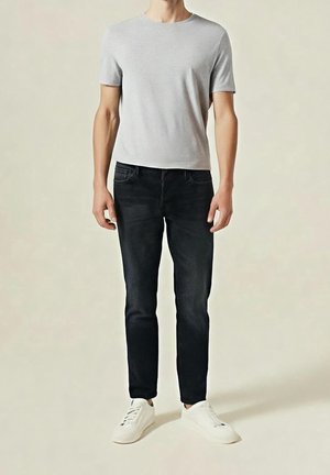 Mann steht und trägt ein hellgraues T-Shirt mit kurzen Ärmeln, schwarze enge Jeans und weiße Turnschuhe vor beigem Hintergrund.