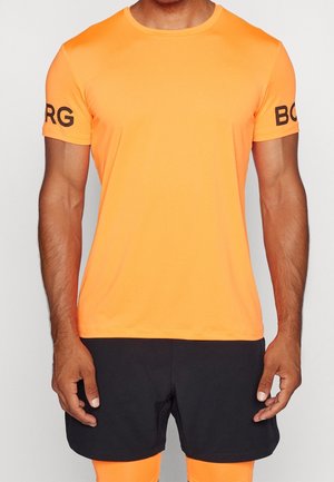 Mand iført en skrigorange kortærmet skjorte med sort tekst på ærmerne og sorte shorts mod en ensfarvet baggrund.