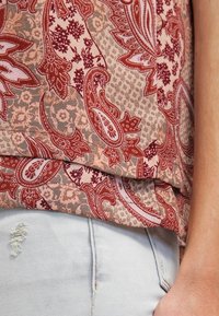 Tessuto rosso con motivo paisley su sfondo beige chiaro e grigio. Presenta un design fluido e un orlo a strati. Indossato con jeans azzurri strappati.