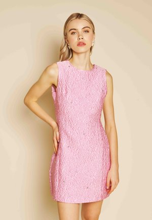 Blondinė moteris, vilkinti rožinės spalvos tekstūruotą mini suknelę be rankovių su mažais papuošimais, pozuoja viena ranka dedama ant klubų prie smėlio spalvos fono.