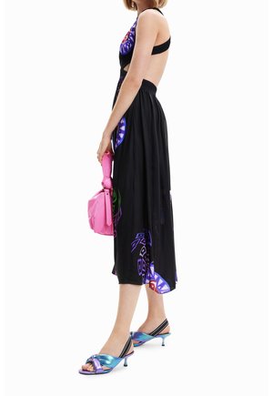 Femme portant une robe noire dos nu avec des motifs colorés, tenant un sac à main rose et portant des sandales à talons ouvertes bleu métallisé.