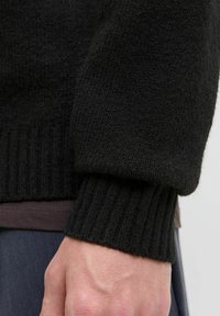 Svart strikket genser med ribbede mansjetter, med en myk tekstur. Nærbilde viser en hånd som hviler på stoffet med et subtilt grått underlag.