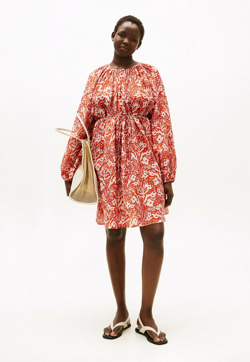 Rotes und weißes Kleid mit Blumenmuster, langen Ärmeln, tailliertem Bund und lockerem Schnitt. Kombiniert mit einer beigefarbenen geflochtenen Tasche und schwarzen Sandalen.