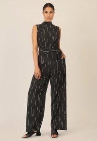 Jumpsuit nero senza maniche con strisce verticali bianche, con cintura in vita, design a gamba ampia, texture liscia e sandali neri aperti sulla punta.