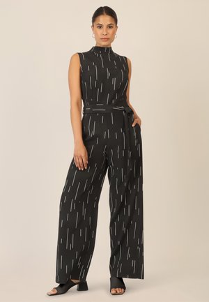Schwarzer ärmelloser Jumpsuit mit weißen vertikalen Streifen, tailliert mit einem Gürtel, weit geschnittene Beine, glatte Textur und flache schwarze offene Schuhe.