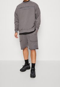 Szara bluza z napisem "CARLO COLUCCI Milano 1978", zestawiona z pasującymi szarymi szortami z dwoma przednimi kieszeniami i detalem logo. Czarne sneakersy.
