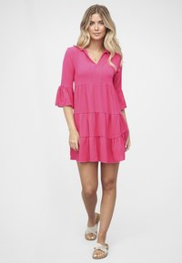 Vaaleanpunainen mekko, jossa on V-aukko, neljännespituiset kellohihat ja porrastettu malli. Sileä kangas, rento istuvuus, polvipituinen, tasaiset sandaalit.