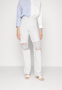 Calças de ganga brancas de cintura alta com detalhes rasgados nos joelhos. Combinadas com uma camisa de botões azul e branca com riscas. Textura suave.