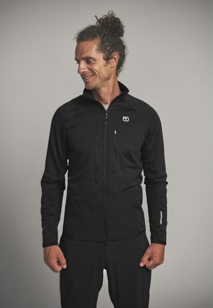 Man with curly hair tied up wears black zip-up jacket and pants, smiling while looking to his left against gray background.