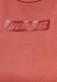 Gros plan sur un tissu texturé brun rougeâtre avec un patch rectangulaire rouge brillant portant le nom de la marque HUMMEL en lettres majuscules grasses.