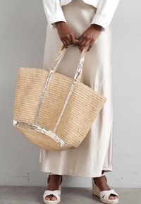 Bolsa de mano de paja tejida con acentos metálicos plateados. Forma grande y redonda con asas dobles. Fondo con un atuendo de tonos neutros.