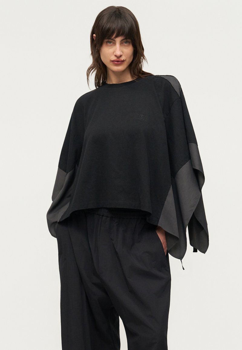 Femme portant un haut noir ample avec de larges manches drapées et un pantalon noir ample assorti, debout avec une main dans la poche.